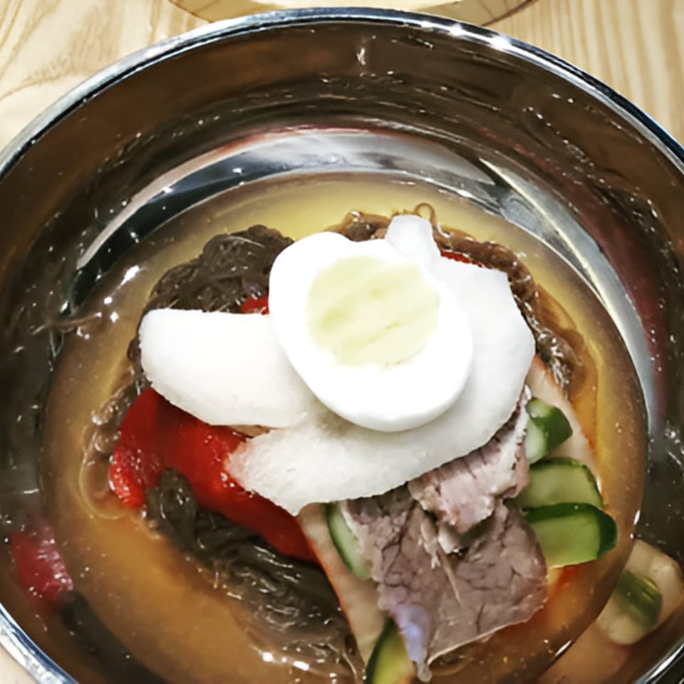 A close-up of Garionban's Korean cold noodles (naengmyeon) with beef, egg, and vegetables in a metal bowl.